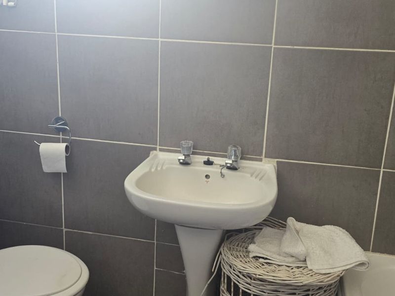 A functional bathroom featuring grey wall tiles, a pedestal basin, a standard toilet, and a bathtub with chrome fixtures.