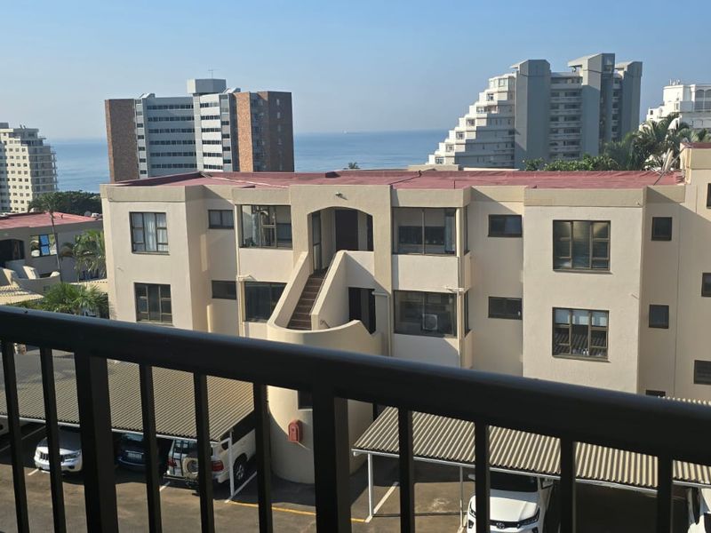 Elevated view overlooking the apartment complex grounds, parking area, and surrounding buildings with a distant ocean horizon.