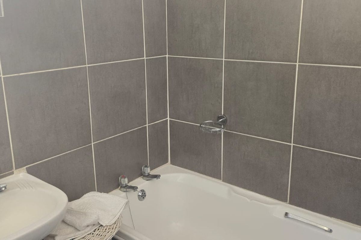 A functional bathroom featuring a white bathtub, pedestal basin, and neutral grey wall tiling throughout.