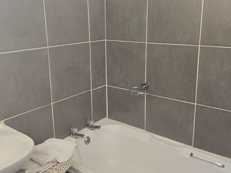 A functional bathroom featuring a white bathtub, pedestal basin, and neutral grey wall tiling throughout.