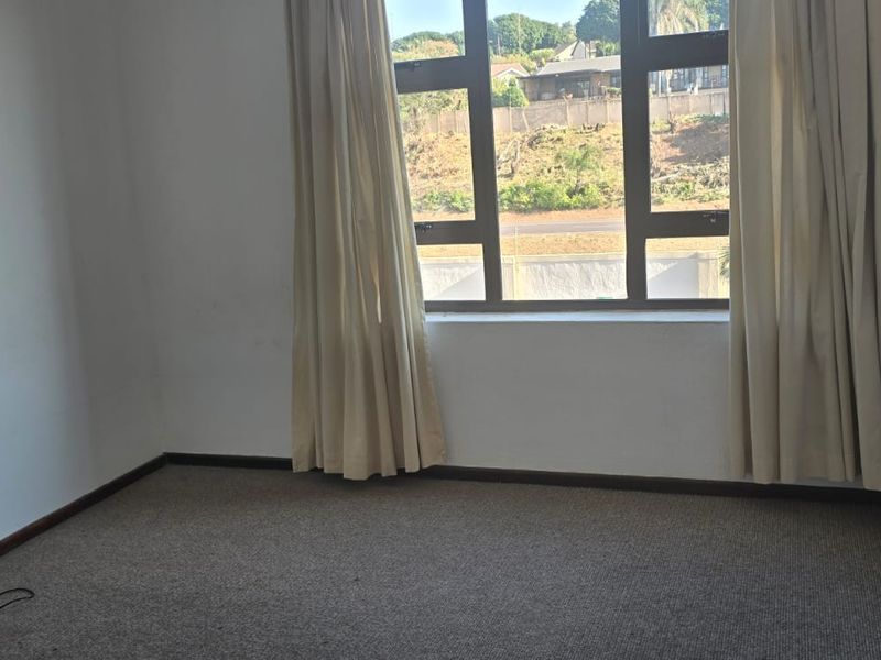 A simple bedroom featuring built-in cupboards, carpeted flooring, and a large window providing natural light and an exterior view.