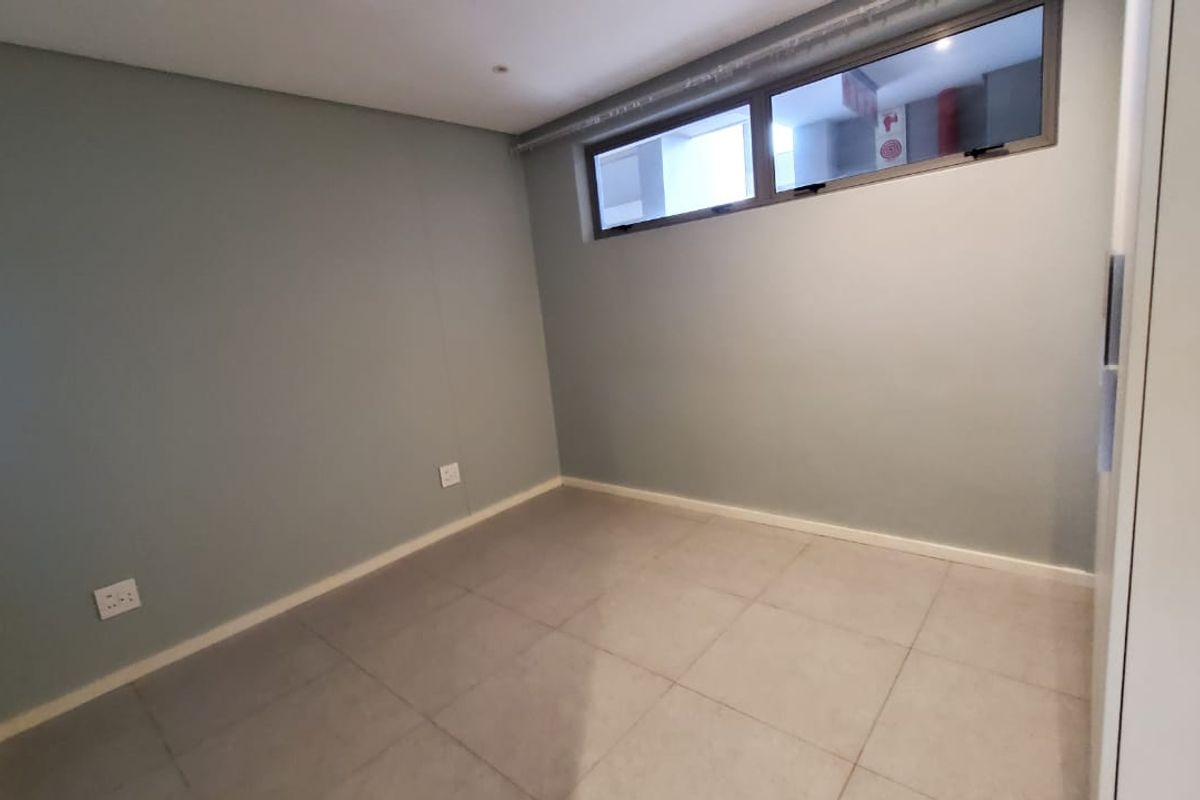 An empty bedroom featuring neutral grey walls, tiled flooring, and a high-level window providing natural light.