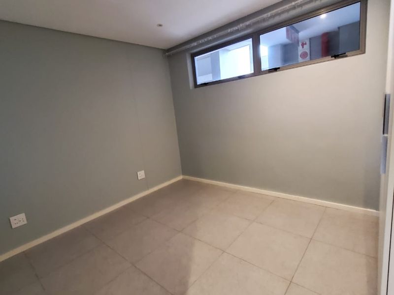 An empty bedroom featuring neutral grey walls, tiled flooring, and a high-level window providing natural light.