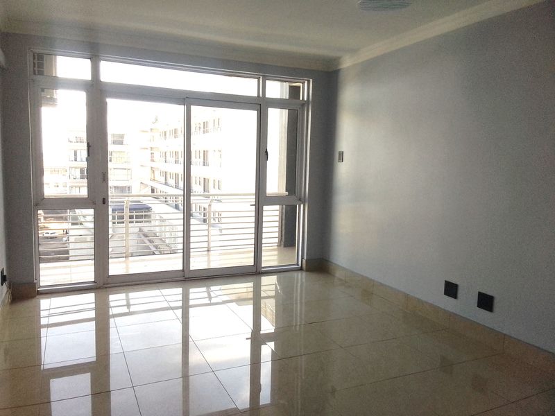 A bright, empty living room featuring polished tile flooring, neutral walls, and a large sliding glass door providing access to the balcony.