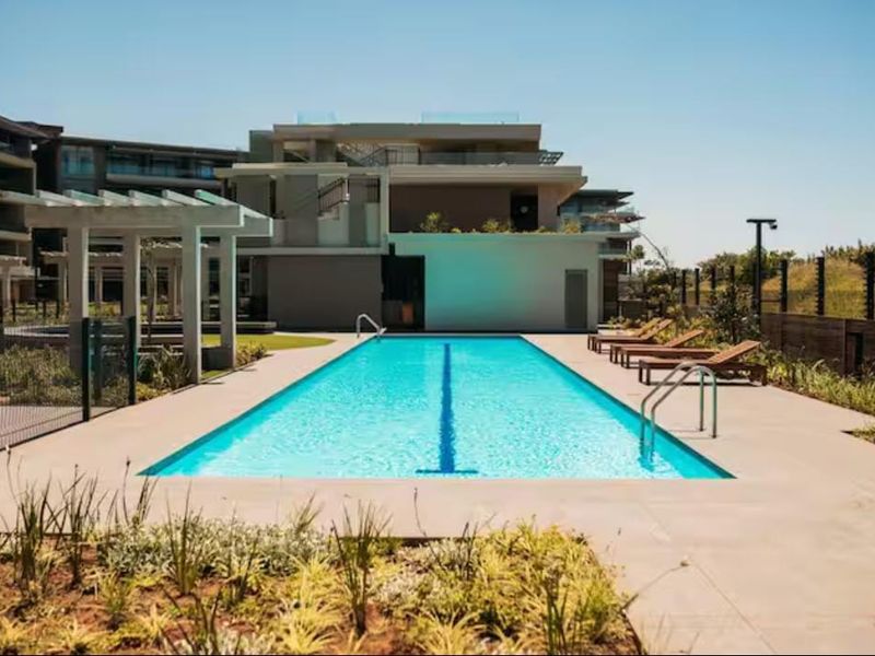 A well-maintained communal swimming pool area featuring a lap pool, paved deck, pergola, and landscaped surroundings within an apartment complex.