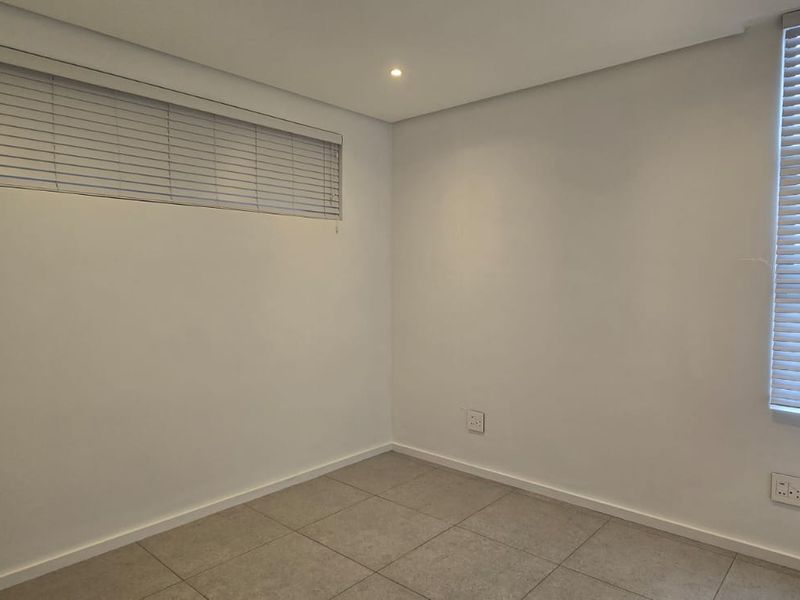 A bright, empty bedroom featuring neutral tiled flooring, recessed ceiling lighting, and fitted venetian blinds on the windows.
