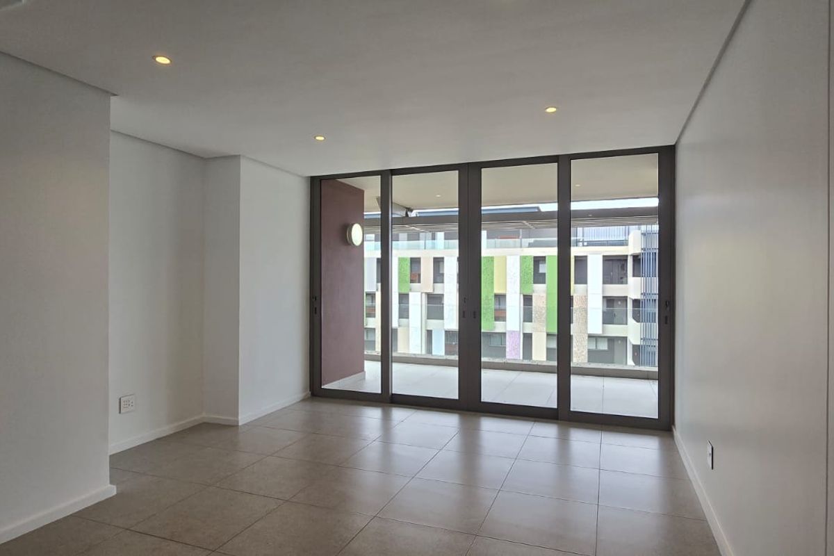 A bright, empty living area featuring neutral tiled floors, recessed lighting, and large sliding glass doors providing access to an outdoor balcony.