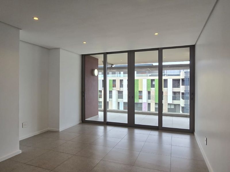 A bright, empty living area featuring neutral tiled floors, recessed lighting, and large sliding glass doors providing access to an outdoor balcony.