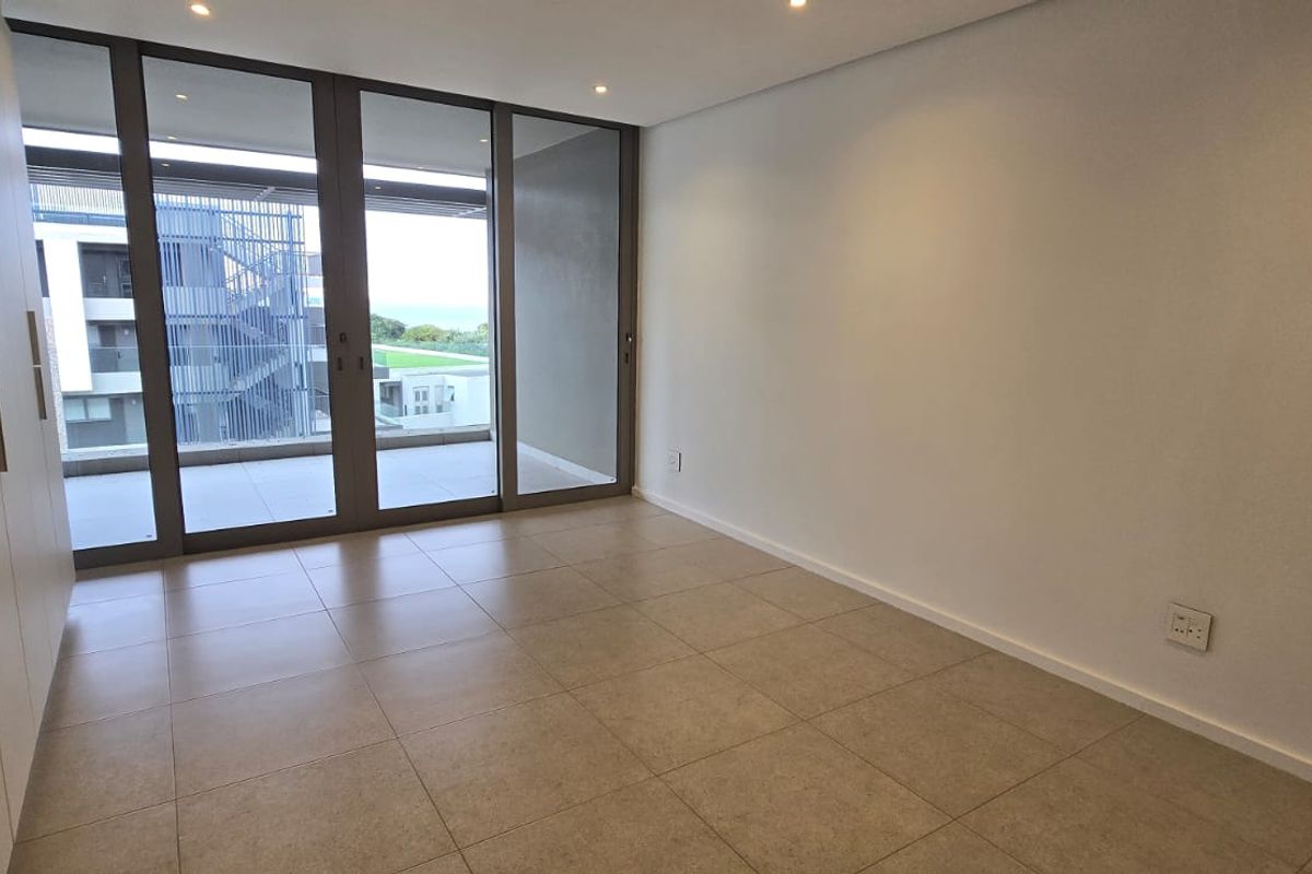 A bright, modern bedroom featuring neutral tiled flooring, ample built-in cupboard space, and large sliding glass doors leading to an outdoor balcony area.