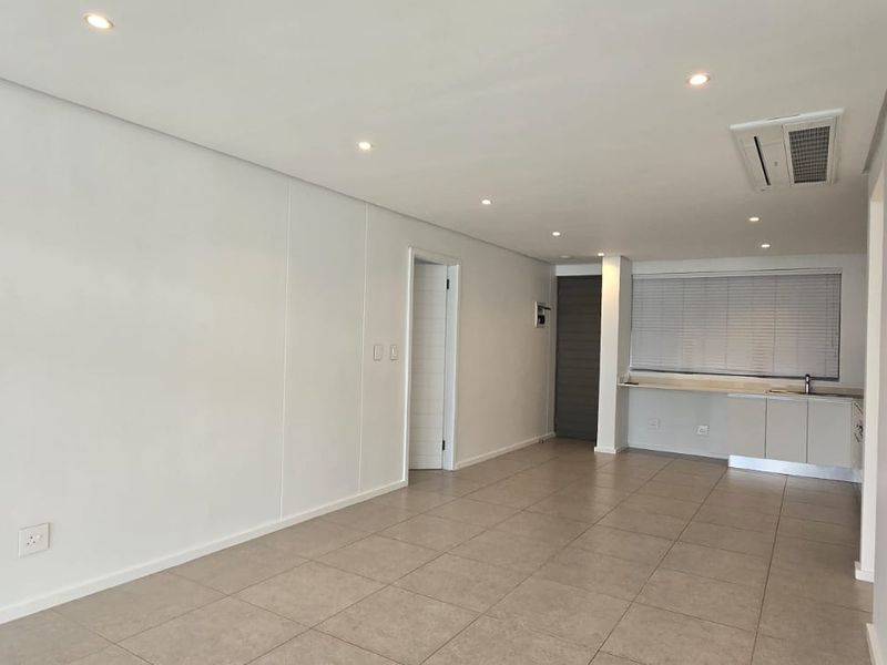 A bright, open-plan living area featuring neutral tiled flooring, recessed lighting, ducted air conditioning, and a compact kitchenette with built-in cabinetry.