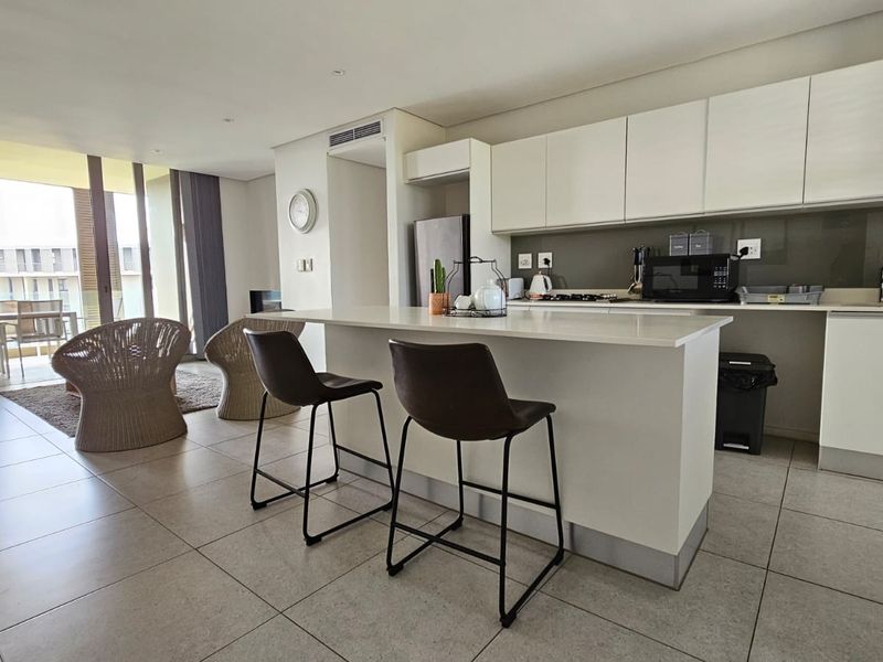 Modern open-plan apartment featuring a sleek kitchen with stone countertops, integrated cabinetry, and tiled flooring, flowing seamlessly into the living area and balcony.