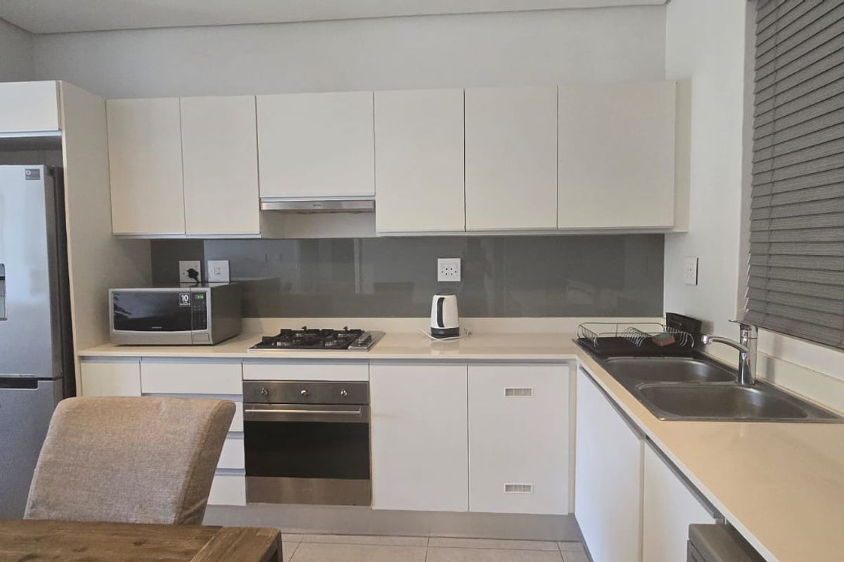 Modern kitchen featuring white cabinetry, stone countertops, glass splashback, gas hob, electric oven, and a double basin sink with venetian blinds.