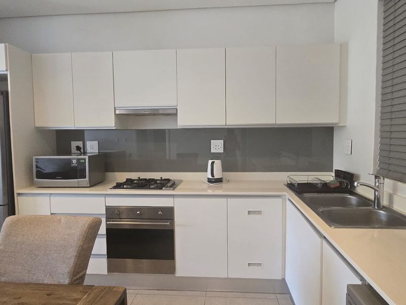 Modern kitchen featuring white cabinetry, stone countertops, glass splashback, gas hob, electric oven, and a double basin sink with venetian blinds.