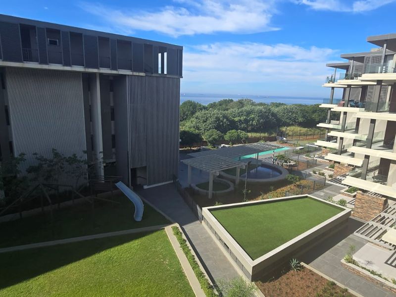 Aerial view of a modern apartment complex featuring landscaped gardens, a swimming pool, paved walkways, and balconies with ocean views.