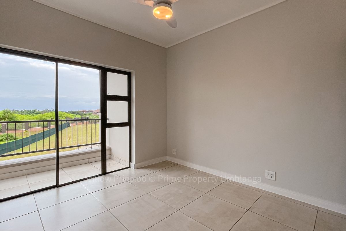 A bright, empty bedroom featuring neutral tiled flooring, a ceiling fan, and a large sliding glass door providing access to a private balcony.