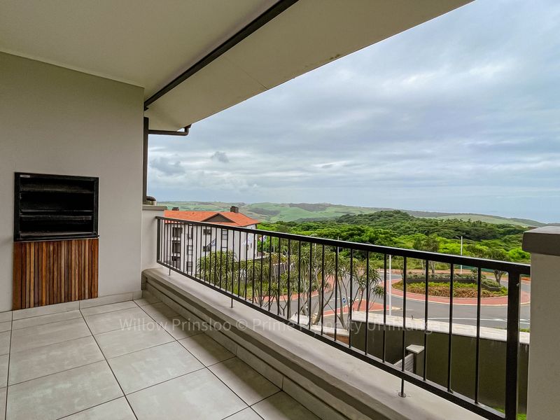 Covered balcony featuring tiled flooring, a built-in braai, and a metal balustrade, offering elevated views of the surrounding landscape and coastal area.