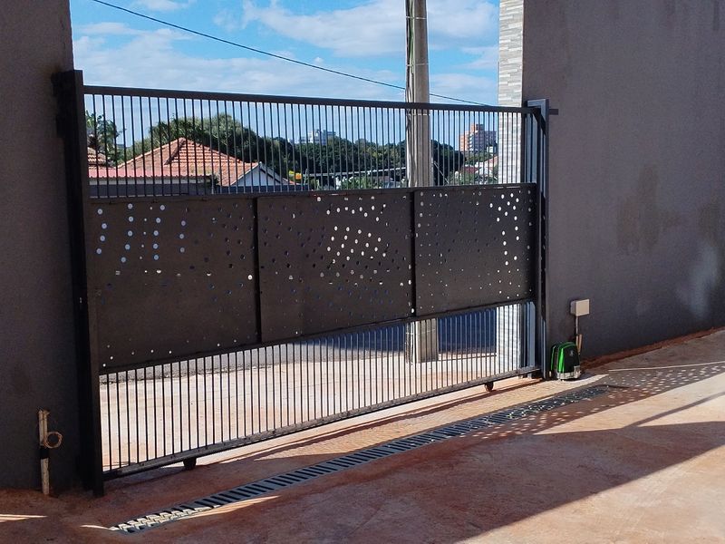 Modern sliding metal driveway gate with decorative perforated panels and a separate pedestrian entrance, set within a secure perimeter wall.