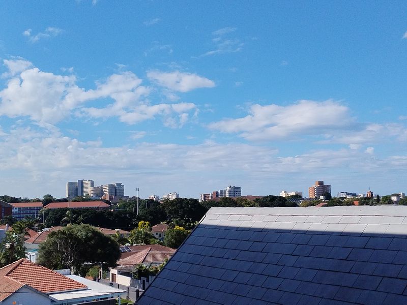 An elevated urban view overlooking surrounding residential rooftops and distant city skyline under a clear blue sky.