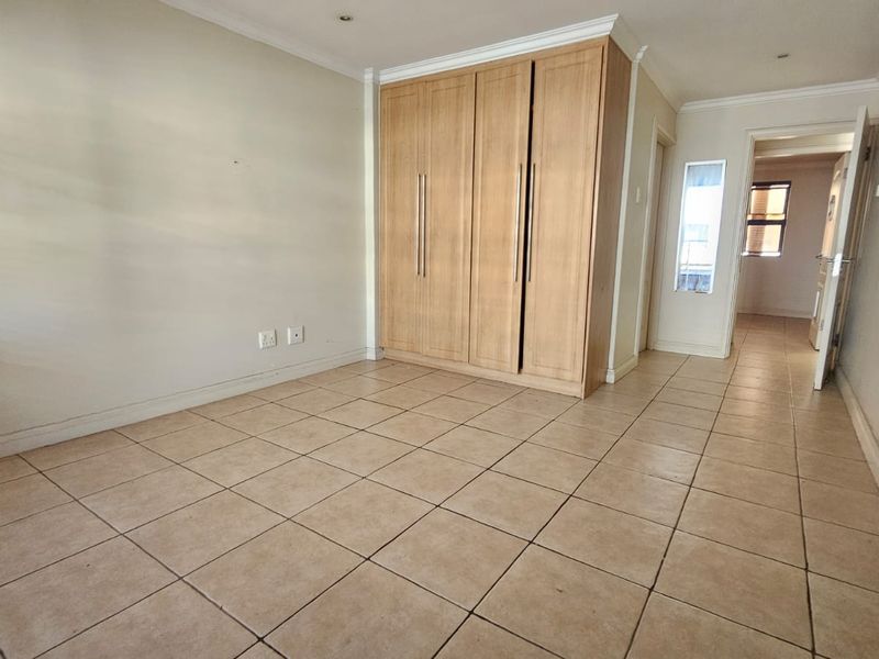 A spacious, empty bedroom featuring neutral tiled flooring, built-in wooden cupboards, and classic ceiling cornicing.