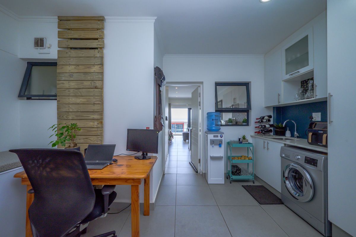 A functional kitchen area featuring white cabinetry, tiled flooring, and a built-in washing machine, with an adjacent workspace and access to the living area.