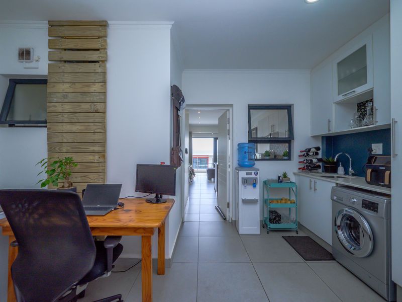 A functional kitchen area featuring white cabinetry, tiled flooring, and a built-in washing machine, with an adjacent workspace and access to the living area.