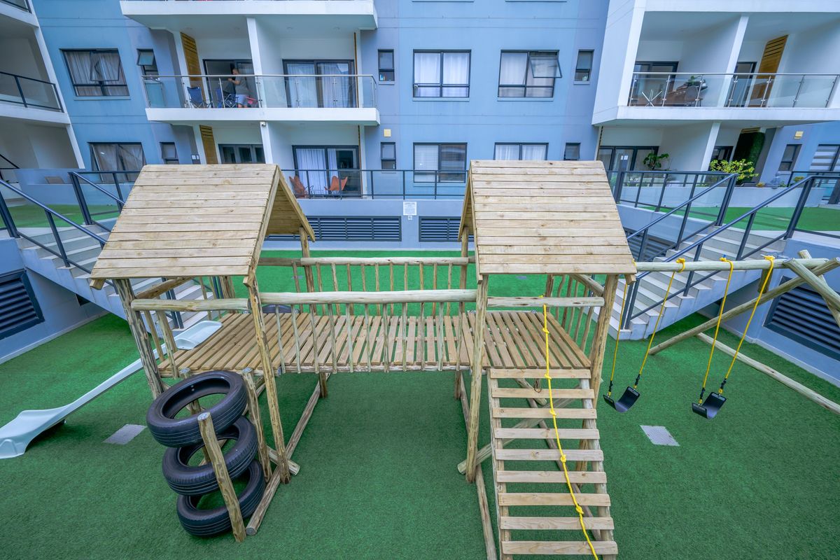 A communal outdoor courtyard area featuring a wooden playground structure with swings, set against the backdrop of a modern apartment building with glass balustrades.