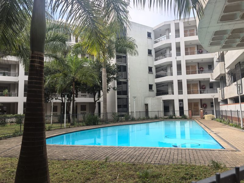 A well-maintained communal swimming pool area set within a multi-story apartment complex, featuring paved decking and landscaped surroundings.