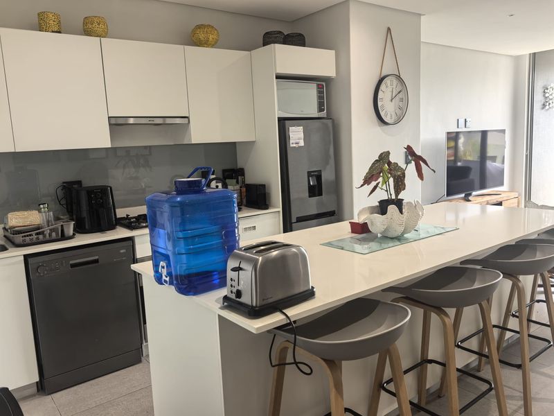 Modern open-plan kitchen featuring sleek white cabinetry, stone countertops, integrated appliances, and a breakfast bar island, seamlessly flowing into the living area.