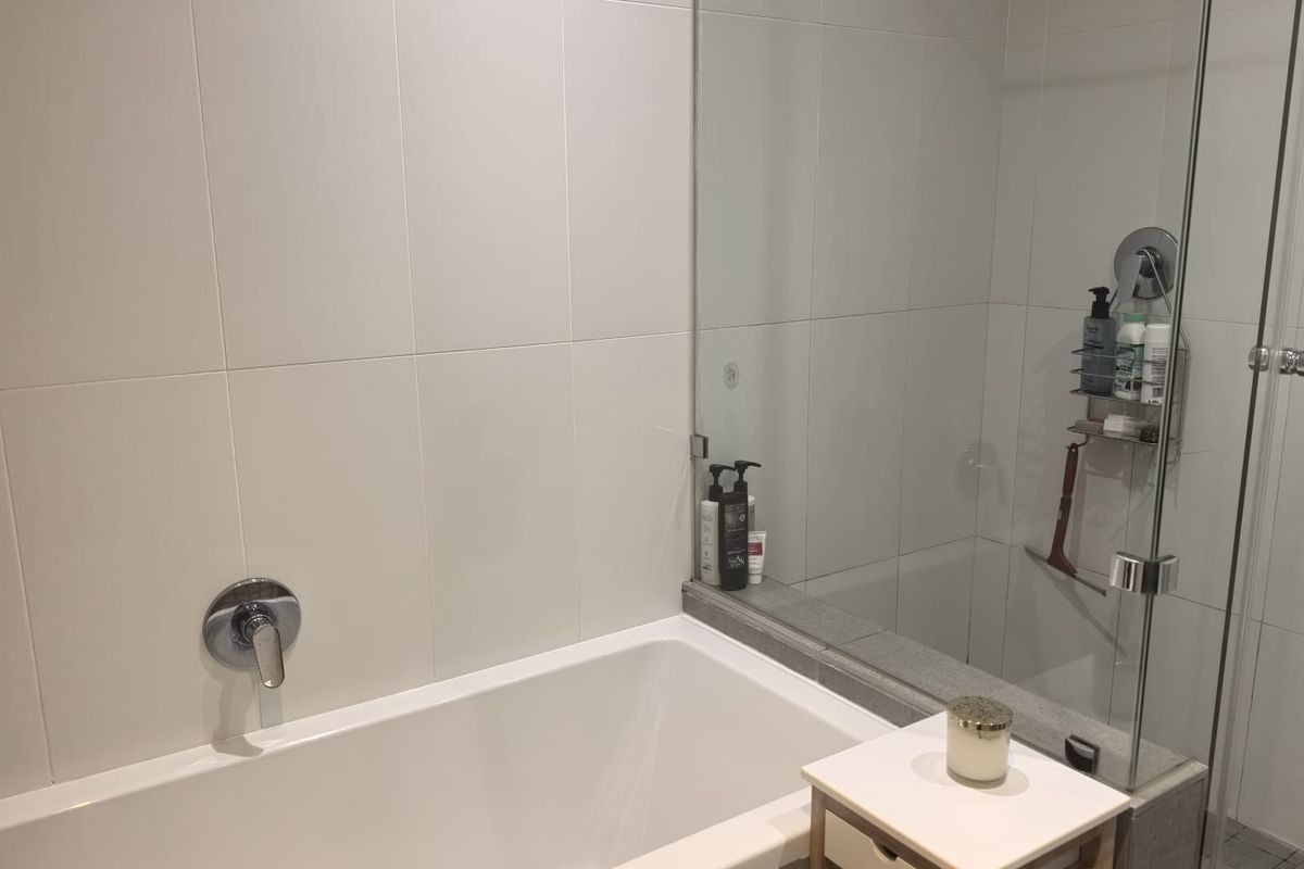 Modern bathroom featuring a built-in bathtub with a glass shower screen and clean, neutral wall tiling.