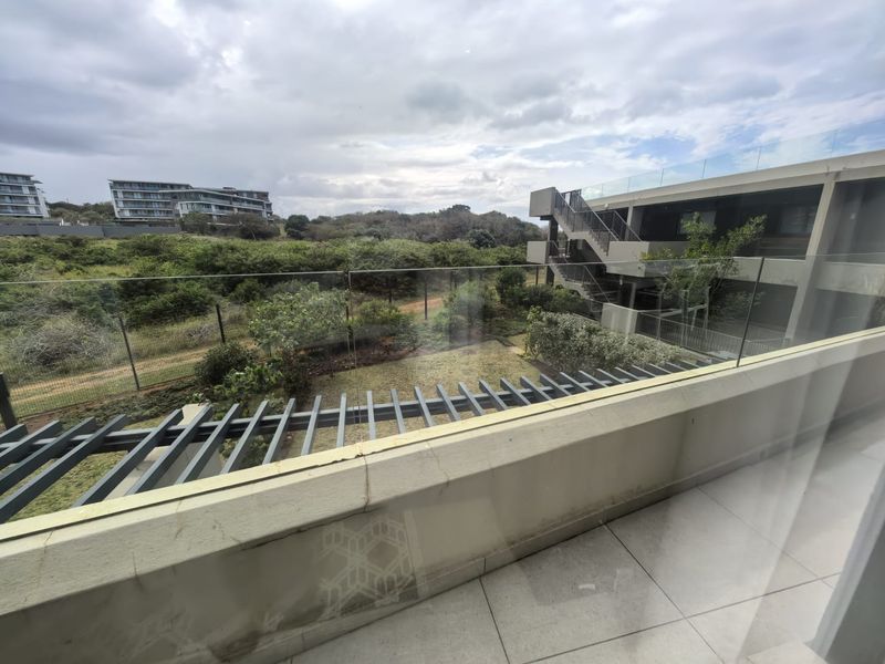A spacious balcony featuring a glass balustrade, tiled flooring, and a pergola, offering views of the surrounding landscape and complex architecture.