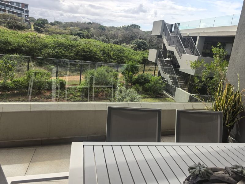 A modern balcony featuring a glass balustrade and tiled flooring, offering an elevated view of the surrounding landscape and adjacent building architecture.