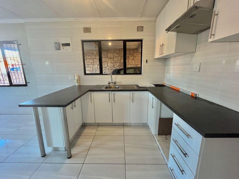 A functional kitchen featuring white cabinetry, dark laminate countertops, a stainless steel sink, and an extractor fan, with tiled flooring and backsplash throughout.