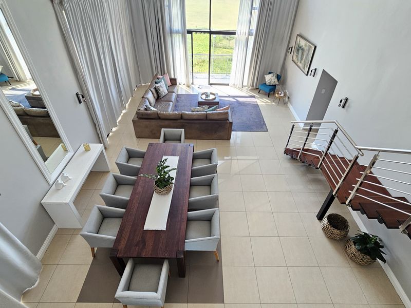 Spacious double-volume open-plan living and dining area featuring large windows, tiled floors, and a modern staircase with stainless steel balustrades.
