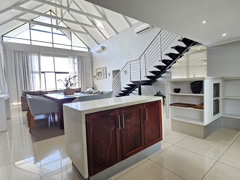 Spacious open-plan living area featuring a vaulted ceiling with exposed trusses, a modern staircase, and a central kitchen island with tiled flooring throughout.