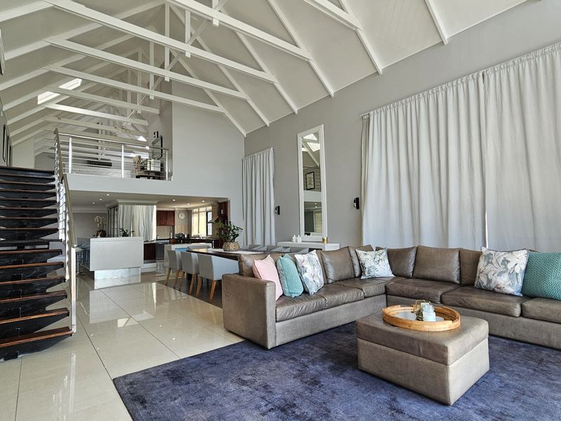 Spacious open-plan living area featuring high vaulted ceilings with exposed trusses, polished tile flooring, and a modern staircase leading to a mezzanine level.