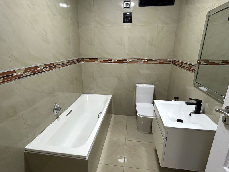 A modern, well-maintained bathroom featuring a bathtub, a sleek vanity with a basin, and neutral wall tiling with a decorative accent border.
