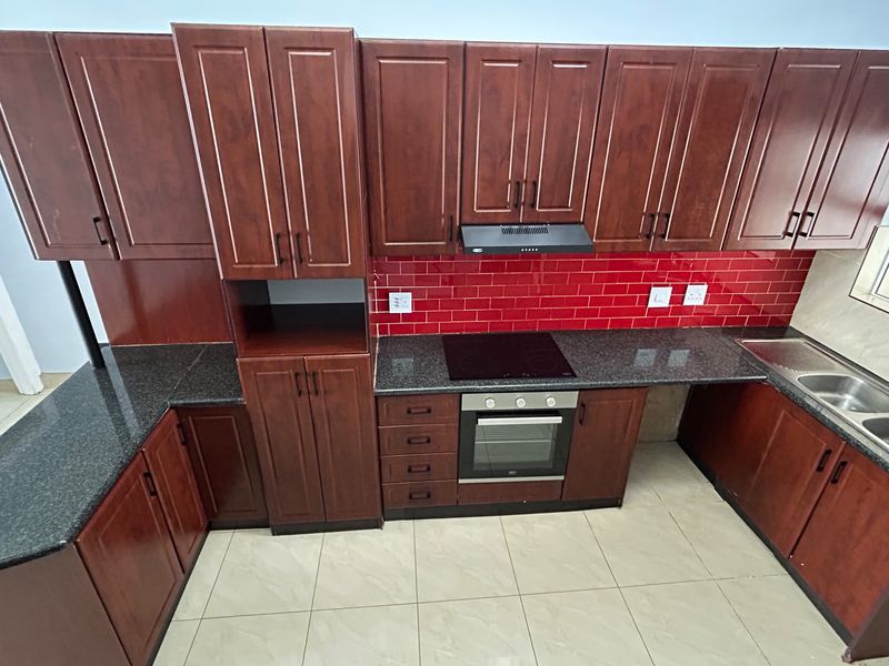 A clean, functional kitchen featuring dark wood cabinetry, granite countertops, a striking red subway tile backsplash, and integrated cooking appliances.