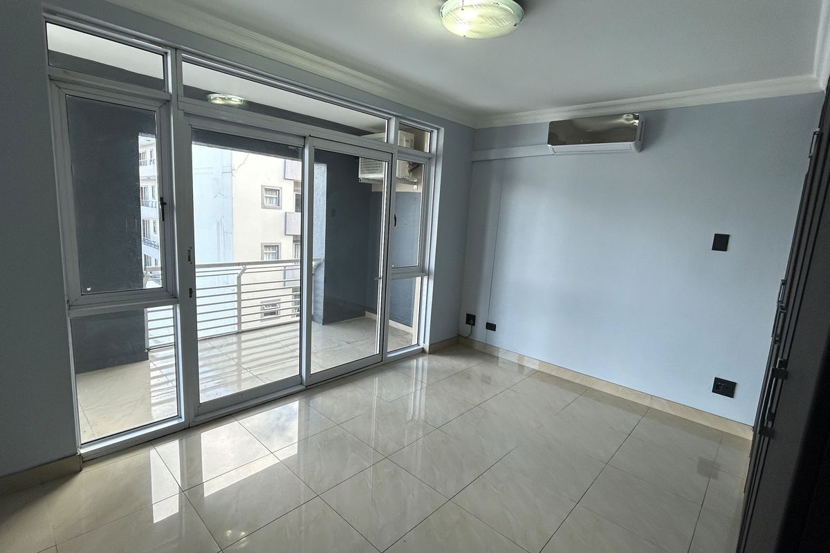 A bright, empty living room featuring large sliding glass doors leading to a balcony, tiled flooring, and a wall-mounted air conditioning unit.