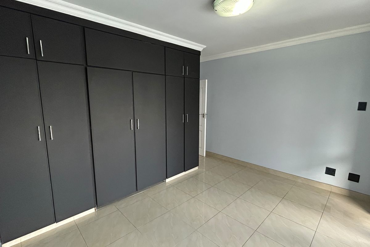 A bright, empty bedroom featuring modern dark built-in cupboards, neutral tiled flooring, and clean wall finishes.