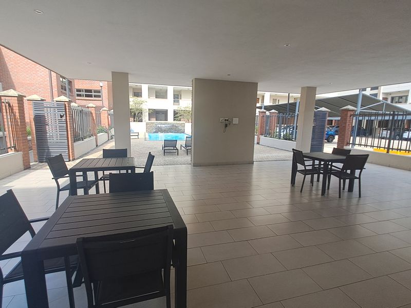 A spacious covered patio area featuring tiled flooring, recessed lighting, and concrete support pillars, overlooking a communal pool and complex grounds.