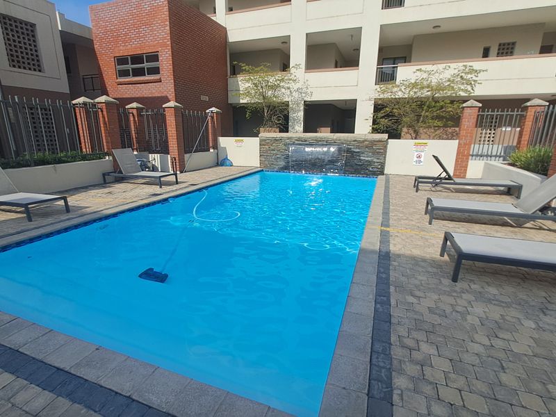 Communal swimming pool area featuring a stone-clad water feature, paved surround, and brick boundary walls within an apartment complex.