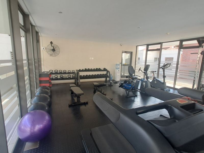 A well-lit apartment gym facility featuring rubberized flooring, floor-to-ceiling windows, and a wall-mounted industrial fan.