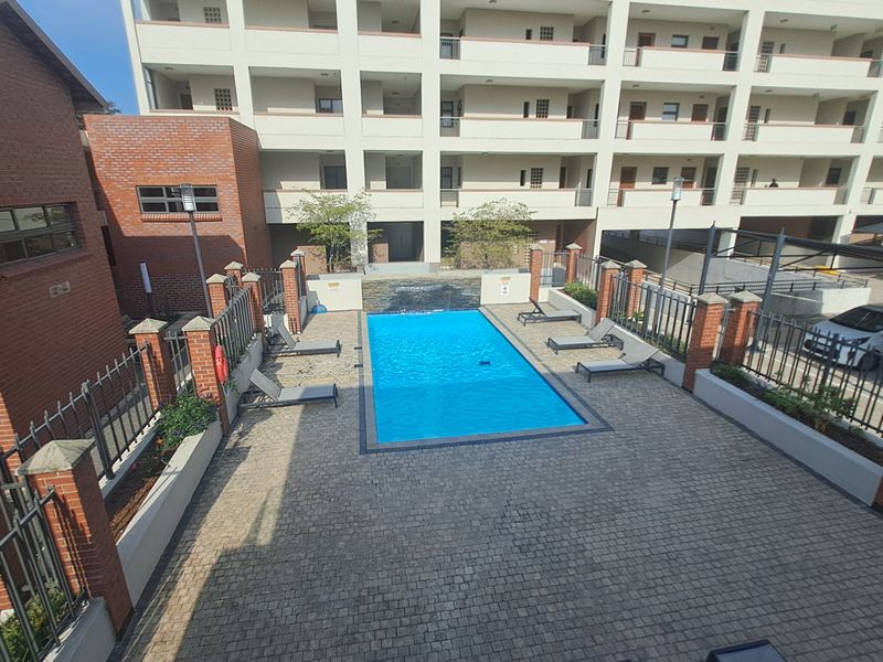 A well-maintained communal swimming pool area featuring a paved deck, water feature, and secure perimeter fencing within an apartment complex.