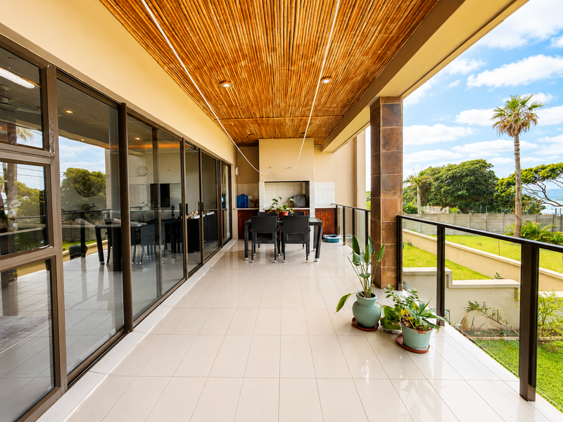 Spacious covered balcony featuring a bamboo ceiling, built-in braai, tiled flooring, and glass balustrades, offering a seamless transition to the outdoor garden area.