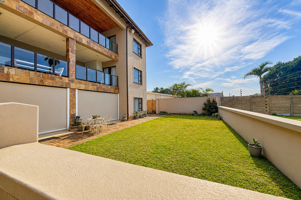 Rear view of the apartment building featuring a well-maintained lawn, paved patio area with retractable blinds, and modern glass balustrades on the upper levels.