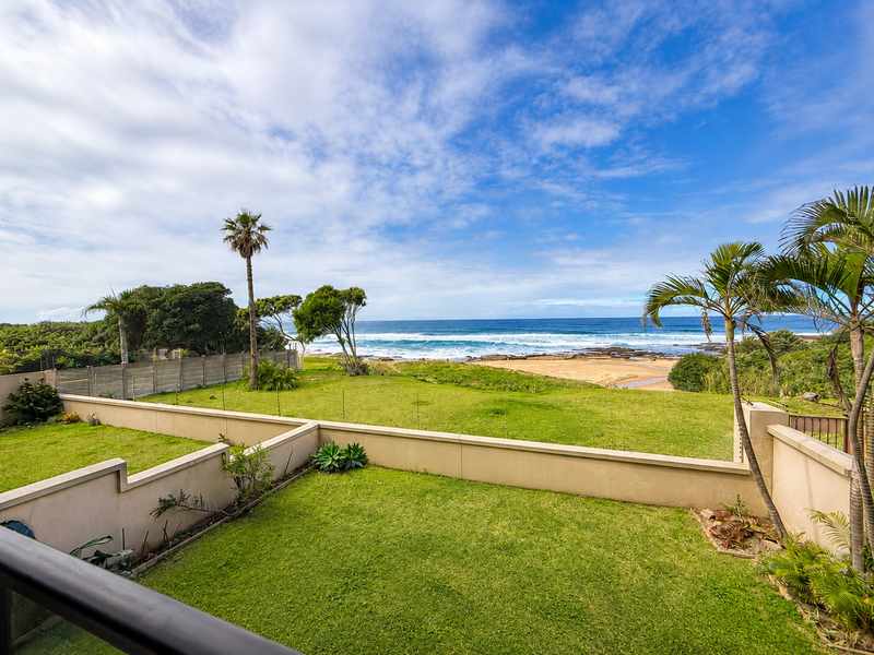 A scenic elevated view overlooking a private lawn, boundary wall with electric fencing, and direct access to the beach and ocean.