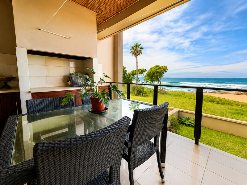 Covered patio featuring a built-in braai, tiled flooring, and a glass balustrade, offering expansive ocean and garden views.