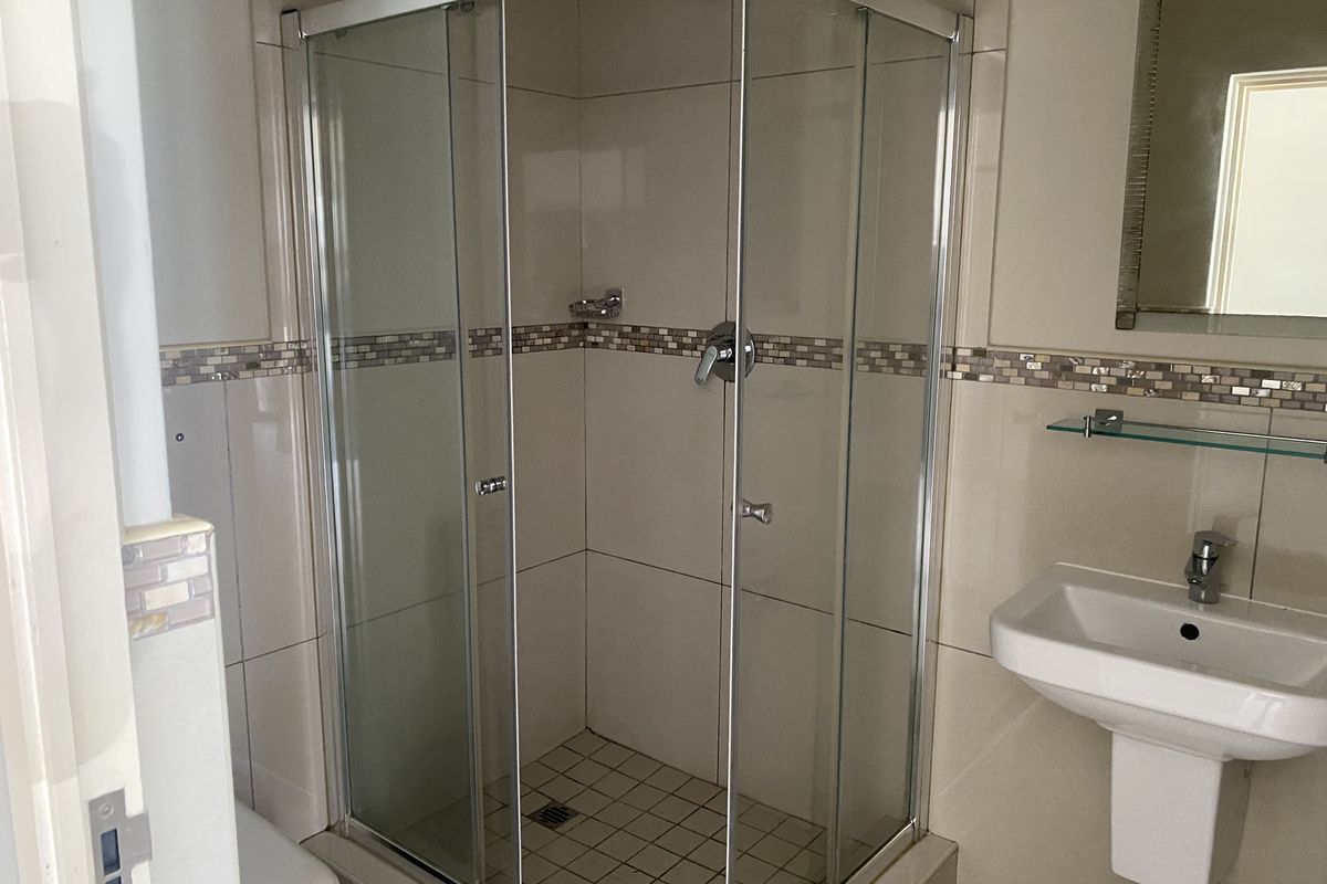 A functional bathroom featuring a corner shower enclosure, pedestal basin, and neutral wall tiling with a decorative mosaic border.