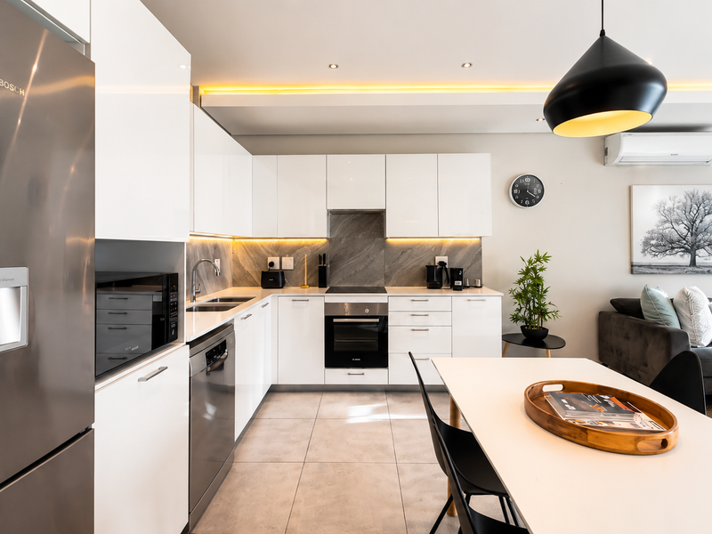 Modern open-plan kitchen and living area featuring sleek white cabinetry, stone countertops, integrated appliances, and contemporary recessed and cove lighting.