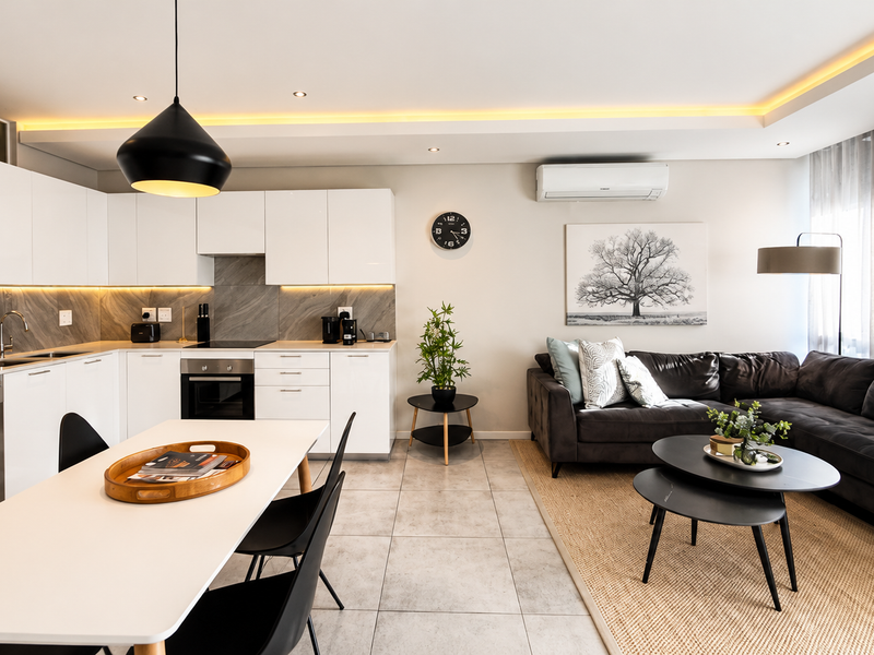 Modern open-plan living area featuring a sleek kitchen with white cabinetry, stone-look backsplash, integrated appliances, and a comfortable lounge space with tiled flooring.
